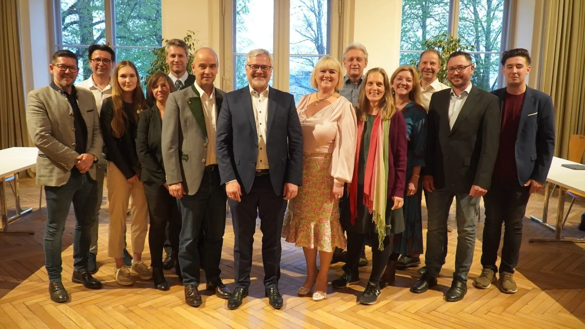 Eine Gruppe von Menschen in formeller Kleidung steht zusammen in einem Raum mit großen Fenstern. Sie lächeln und posieren für ein Foto. Hinter ihnen sind Fenster mit Bäumen draußen.