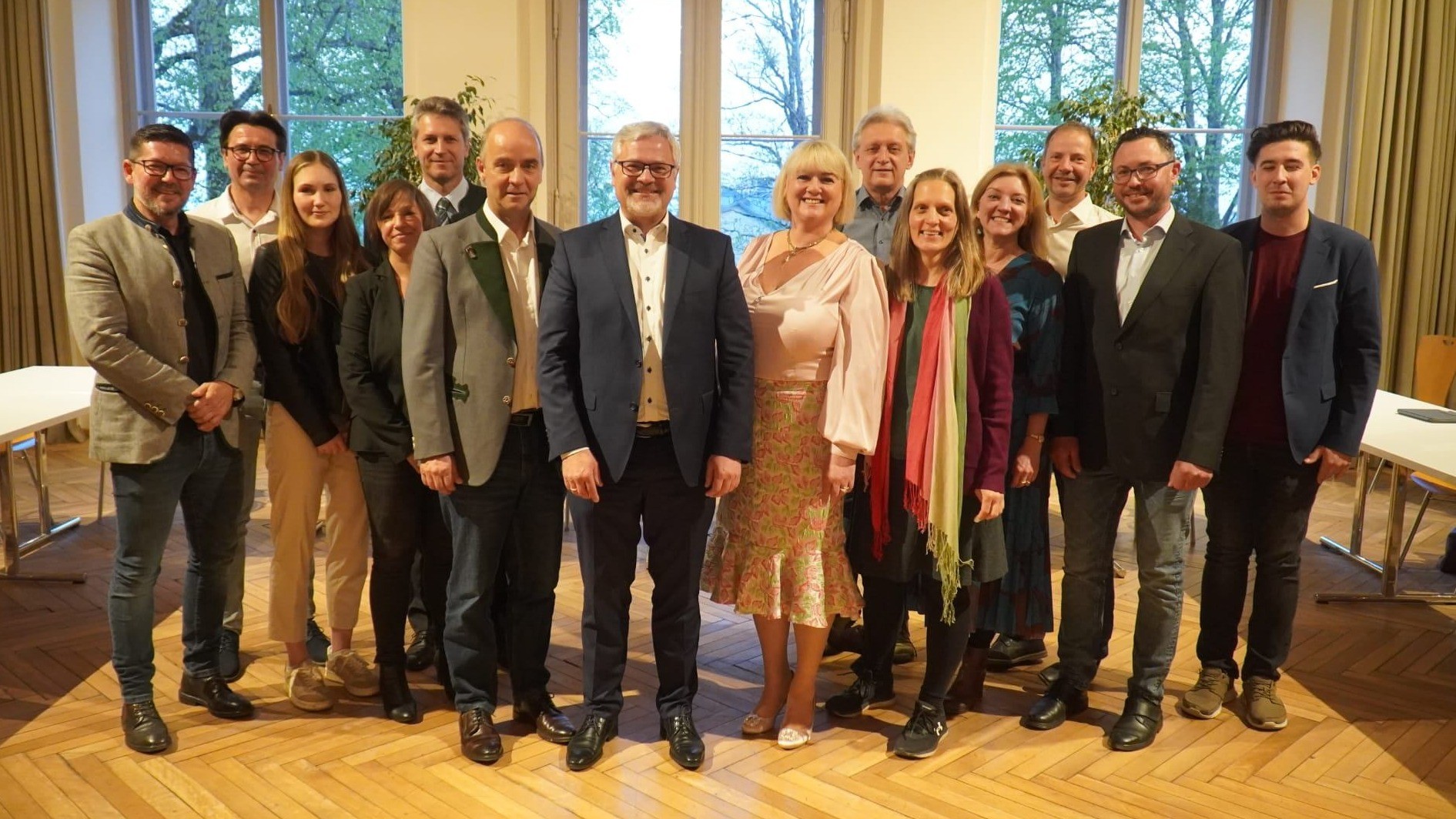 Eine Gruppe von Menschen in formeller Kleidung steht zusammen in einem Raum mit großen Fenstern. Sie lächeln und posieren für ein Foto. Hinter ihnen sind Fenster mit Bäumen draußen.