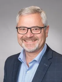 Ein Mann mit weißem Haar und Bart, der eine Brille und ein blaues Hemd trägt, lächelt und scheint für ein Foto zu posieren.