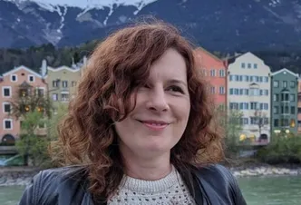 Eine lächelnde Frau mit lockigem rotem Haar steht vor bunten Gebäuden und einem Fluss. Im Hintergrund befinden sich Berge.