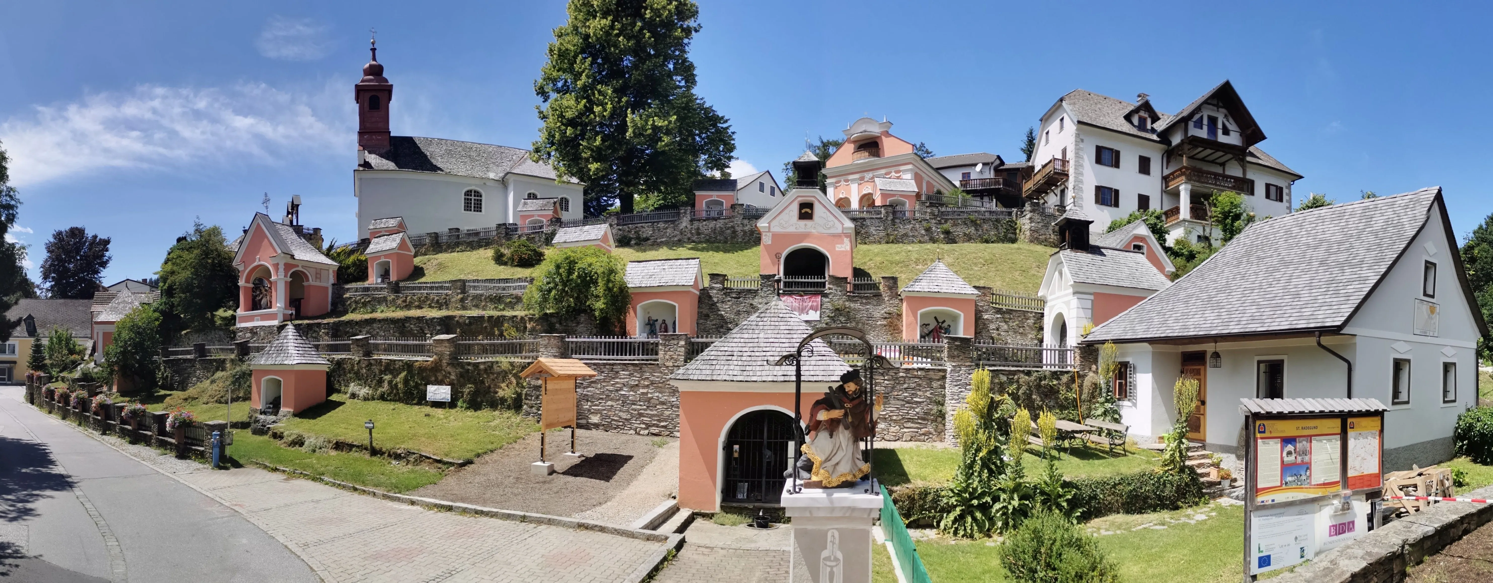 Eine rosa Kapelle auf einem Hügel, umgeben von Steinmauern, mit einer Statue eines Mannes in einem Gewand. Die Kapelle hat einen Glockenturm und ein Kreuz auf der Spitze. Bäume und Pflanzen sind in der Nähe.