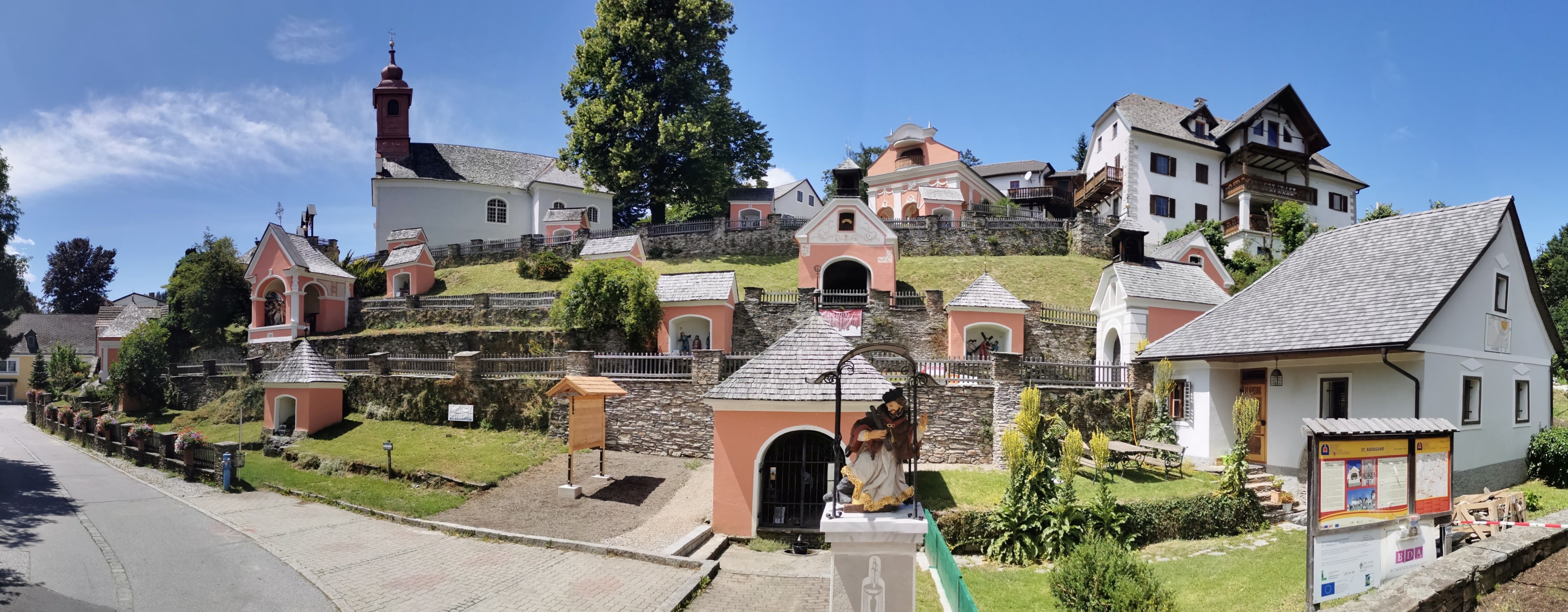 Eine rosa Kapelle auf einem Hügel, umgeben von Steinmauern, mit einer Statue eines Mannes in einem Gewand. Die Kapelle hat einen Glockenturm und ein Kreuz auf der Spitze. Bäume und Pflanzen sind in der Nähe.