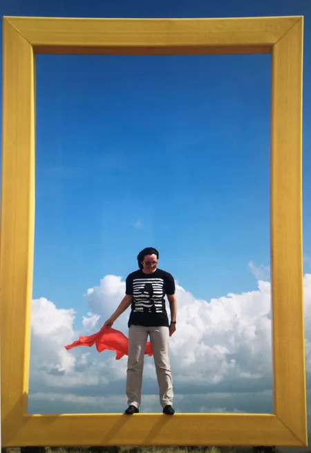 Ein Mann steht vor einem Himmelshintergrund mit Wolken. Er hält ein rotes Tuch und trägt ein schwarzes T-Shirt mit weißen Streifen.