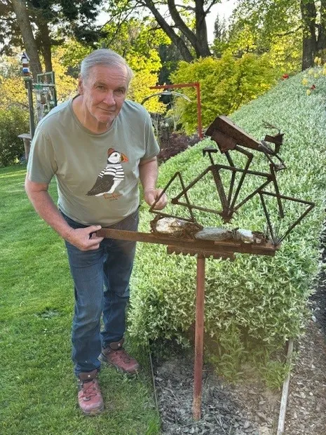 Ein älterer Mann steht in einem Garten und hält eine Metallvogelskulptur. Er trägt ein graues Shirt mit einem Puffin-Aufdruck. Der Garten hat grünen Rasen und Sträucher.