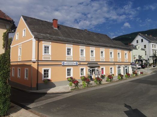 Ein beigefarbenes Gebäude mit grauem Dach und Schornstein, mit weißen Fenstern und einem Schild, das Gasthaus Kirchenwirt besagt. Es ist mit Blumenkästen und Pflanzen vor dem Gebäude geschmückt.