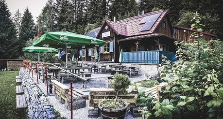 Eine rustikale Außengastronomie mit Holztischen und Bänken unter grünen Sonnenschirmen, einem Haus mit Solarmodulen auf dem Dach und einer Einfriedung im Vordergrund.