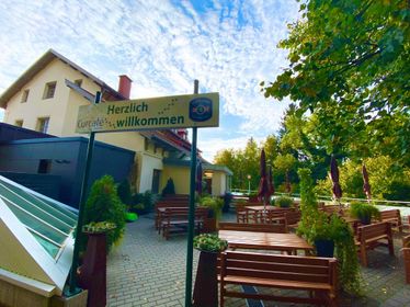 Ein Außenbereich mit Holztischen und -bänken, einem gelben Schild mit der Aufschrift 'Herzlich willkommen' und einem blauen Himmel mit Wolken.