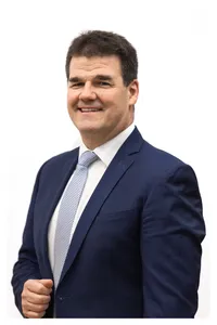 Ein Mann in einem dunkelblauen Anzug mit weißem Hemd und gemusterter Krawatte lächelt und posiert für ein Foto.