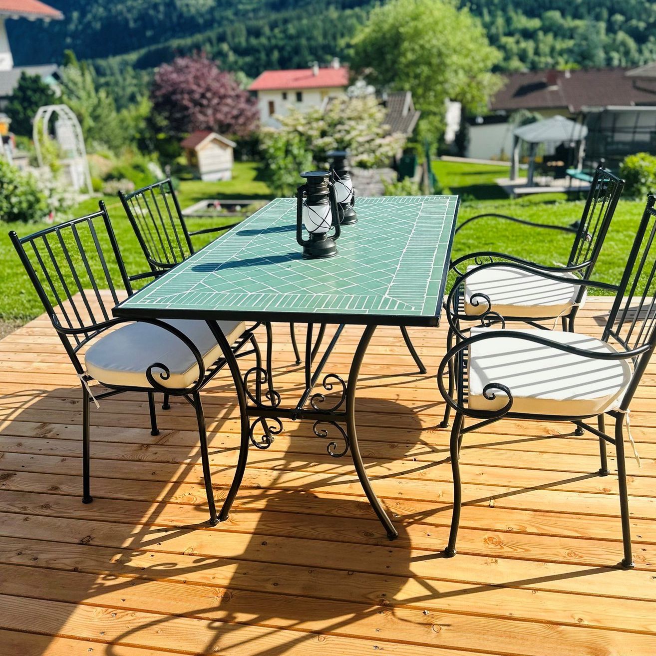 Eine Holzterrasse mit einem grün gefliesten Tisch und vier schwarzen Metallstühlen mit weißen Kissen. Zwei Laternen stehen auf dem Tisch. Die Terrasse ist von einem Rasenbereich mit Bäumen und Häusern im Hintergrund umgeben.