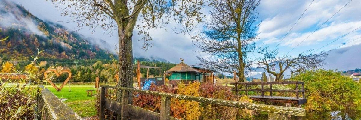 Ein kleiner Holzzaun umgibt einen Garten mit einem grünen Pavillon, einer blauen Rutsche und einer Holzbank. Der Garten ist von Bäumen, Büschen und einem Grasfeld umgeben. Im Hintergrund befinden sich Berge und ein blauer Himmel mit Wolken.