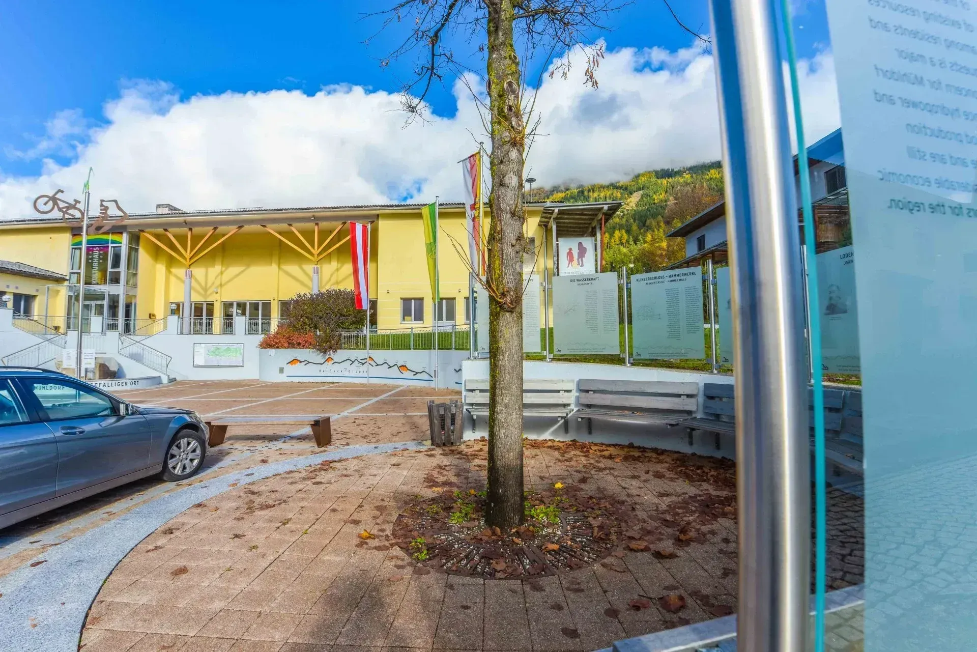 Ein gelbes Gebäude mit Fahnen steht hinter einem Baum und Bänken in einem sonnigen Bereich. Ein Auto ist davor geparkt.