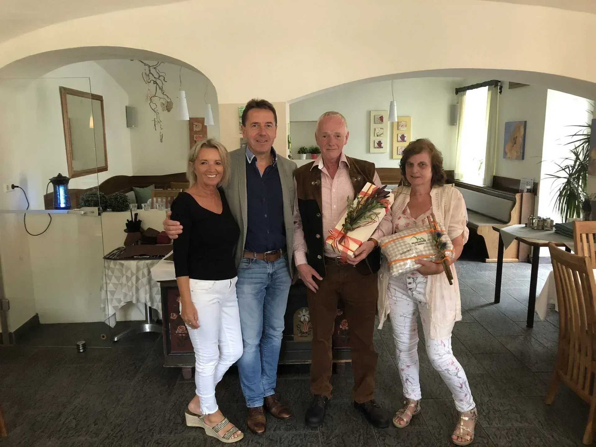 Vier Menschen stehen lächelnd in einem Raum. Ein Mann in einer braunen Jacke hält Blumen und ein Geschenk. Zwei Frauen halten Geschenke und Blumen, und ein weiterer Mann in einem blauen Hemd steht mit seinem Arm um eine Frau.