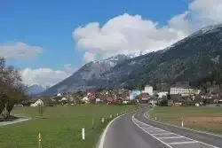 Eine leere Straße kurvt durch ein grünes Tal, mit Häusern und Gebäuden auf der rechten Seite. Berge mit schneebedeckten Gipfeln sind im Hintergrund. Der Himmel ist teilweise bewölkt.