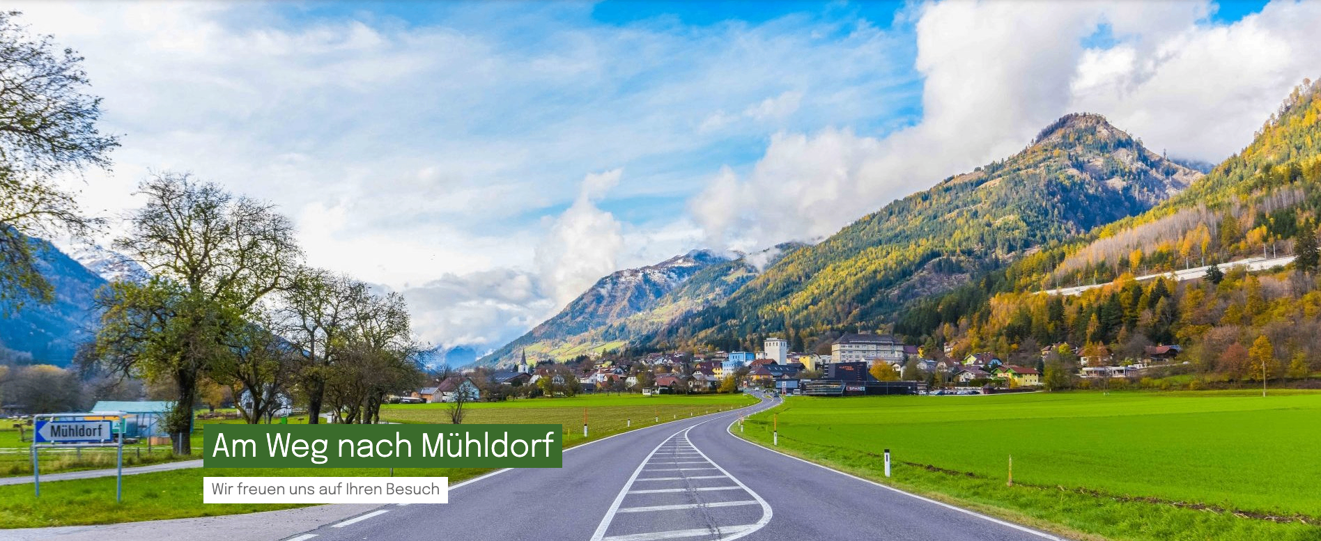 Eine Straße kurvt durch ein üppiges Tal, mit Bergen und einem Dorf in der Ferne. Der Himmel ist teilweise bewölkt.