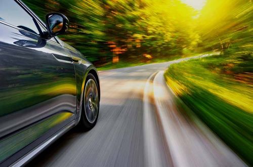 Ein schlankes schwarzes Auto rast mit verschwommenen Bäumen und Sonnenlicht auf einer kurvenreichen Straße.