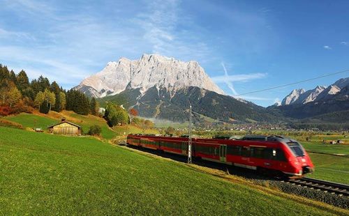 Ein roter Zug fährt auf einer Bahnstrecke durch ein saftiges grünes Tal unter einem blauen Himmel mit Bergen im Hintergrund.