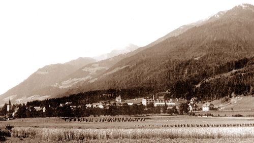 Ein Schwarz-Weiß-Bild zeigt ein Dorf umgeben von Feldern und Bergen. Menschen ernten im Vordergrund Feldfrüchte.