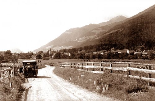 Ein altes Foto zeigt ein Auto, das auf einer unbefestigten Straße mit einem Zaun an einer Seite fährt, umgeben von einer ländlichen Landschaft mit Bergen im Hintergrund.