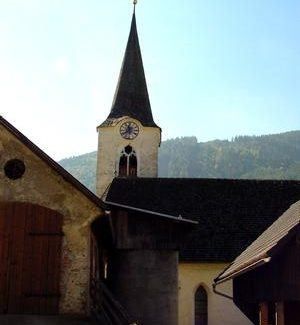 Eine Kirche mit einem Kirchturm und einer Uhr, vor einem Bergrücken mit blauem Himmel.