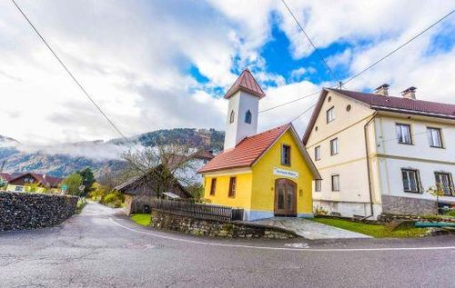 Ein kleines gelbes Haus mit rotem Dach und Turm steht an einer Straße mit Bergen im Hintergrund.