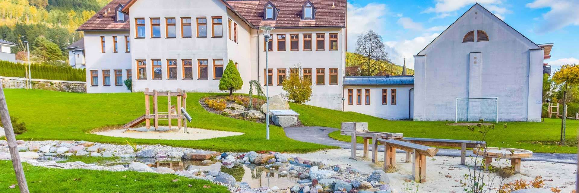 Ein großes weißes Gebäude mit mehreren Fenstern und einem braunen Dach. Vor dem Gebäude befindet sich ein kleiner Teich mit Steinen, einer Bank und einer Straßenlampe.