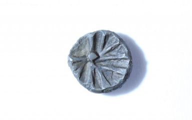 Ein rundes, flaches graues Objekt mit einem radialen Muster und einem kleinen zentralen Buckel, auf einer weißen Oberfläche platziert.