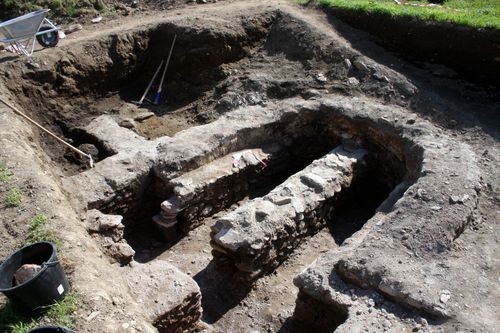 Archäologische Ausgrabungsstätte mit sichtbaren Steinstrukturen und Werkzeugen. Steinmauern und Artefakte deuten auf menschliche Aktivität in der Vergangenheit hin.