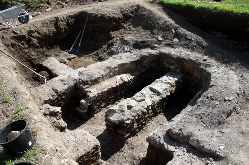 Archäologische Ausgrabungsstätte mit sichtbaren Steinstrukturen und Werkzeugen. Steinmauern und Artefakte deuten auf menschliche Aktivität in der Vergangenheit hin.