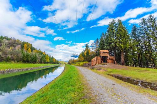 Ein malerischer Weg neben einem Kanal mit einer kleinen Holzstruktur auf der rechten Seite. Der Weg wird von üppigem Gras und Bäumen unter einem blauen Himmel gesäumt.