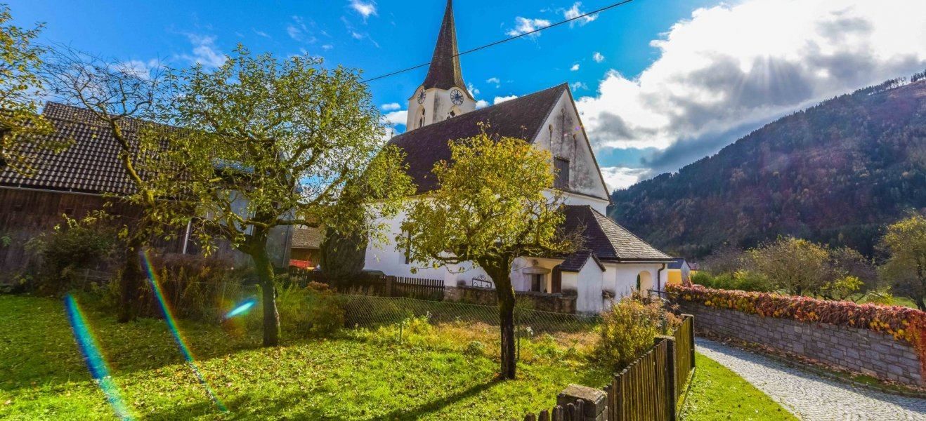 Eine kleine weiße Kirche mit einem Turm und einer Uhr, umgeben von einem Zaun und Bäumen, mit einem Berg im Hintergrund.
