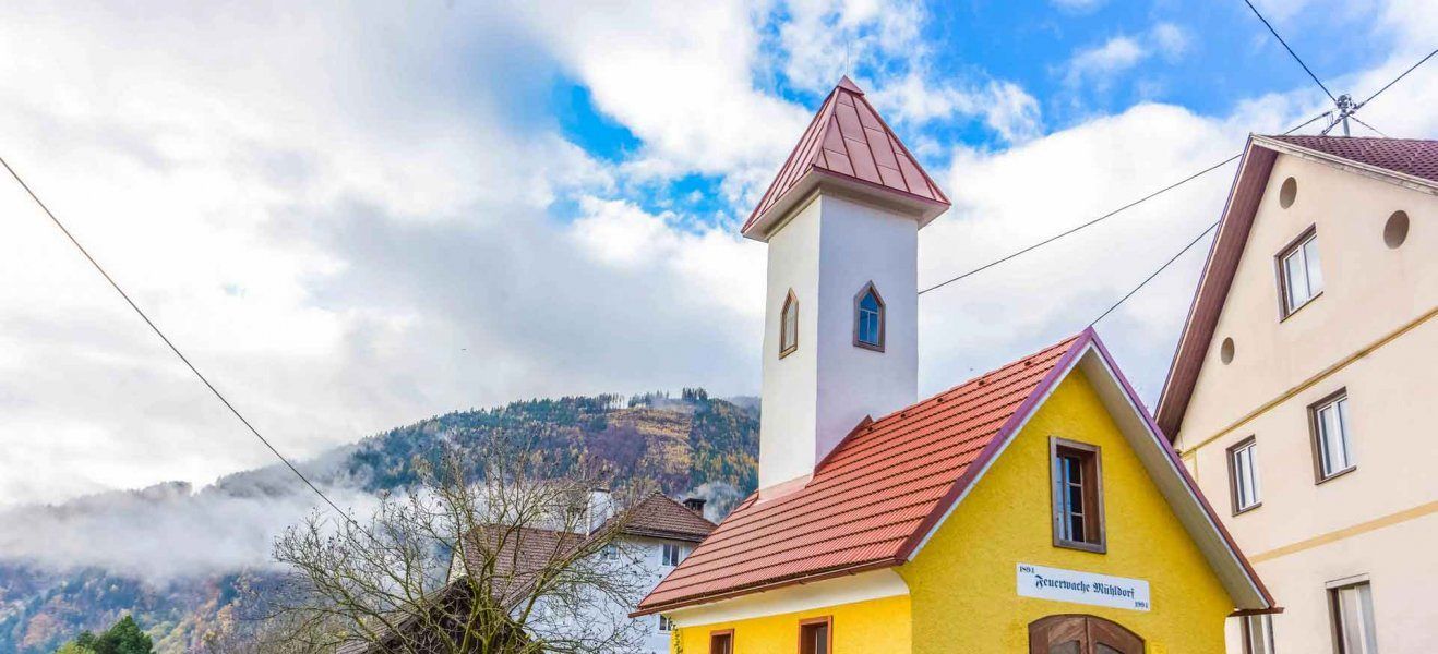 Eine kleine gelbe Kirche mit einem roten Dach und einem Kirchturm steht gegen einen bewölkten Himmel mit einem Berg im Hintergrund.