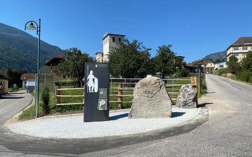 Ein Denkmal mit einer Kriegerfigur steht in der Nähe eines großen Felsens in einem ländlichen Gebiet, mit einer Straße und Häusern im Hintergrund unter einem klaren Himmel.
