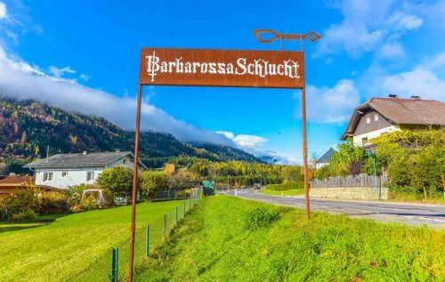 Ein Schild mit der Aufschrift Barbarossaschlucht steht in einem Grasfeld. Im Hintergrund sind eine Straße und Häuser zu sehen.