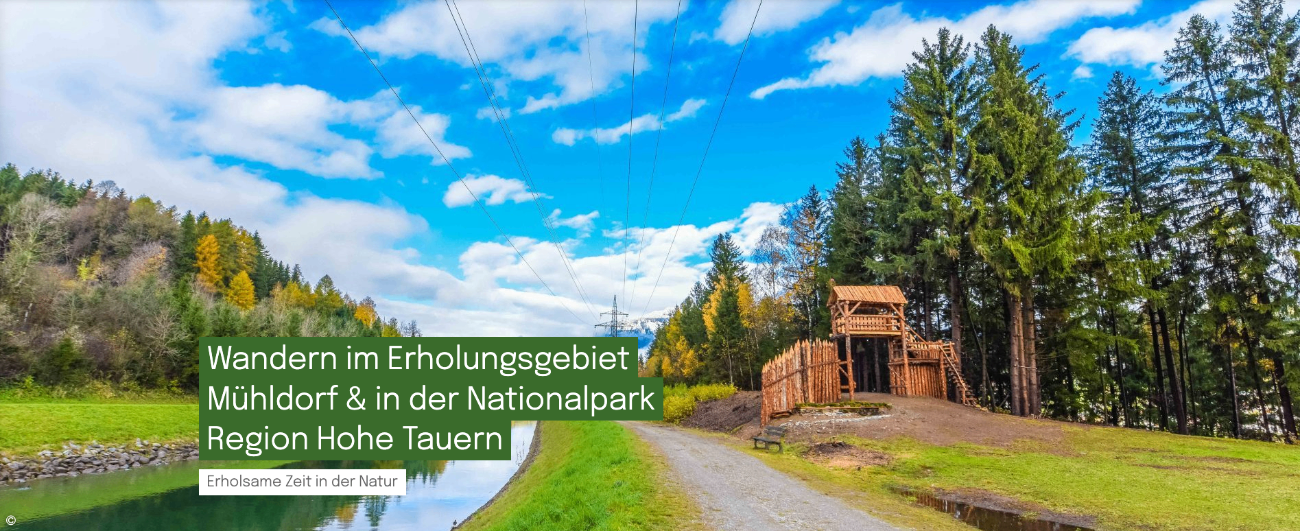 Ein Weg im Nationalpark Tauren führt durch eine Landschaft mit einem See, Bäumen und einer Holzstruktur. Der Himmel ist blau mit verstreuten Wolken.