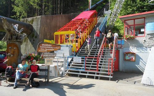 Menschen auf den Stufen einer gelben Standseilbahn am Fuße eines Berges. Zwei Frauen sitzen auf einer Bank auf der linken Seite.
