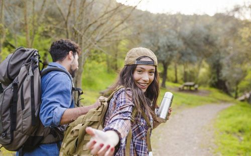 Eine lächelnde Frau in einem karierten Shirt und Hut hält einen Thermos, sie steht mit einem Mann auf einem Pfad in einem bewaldeten Gebiet. Hinter ihnen sind Bäume und eine Picknickbank zu sehen.