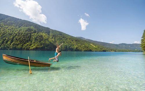 Zwei Jungen springen an einem sonnigen Tag von einem Boot in einen See, mit Bergen im Hintergrund.