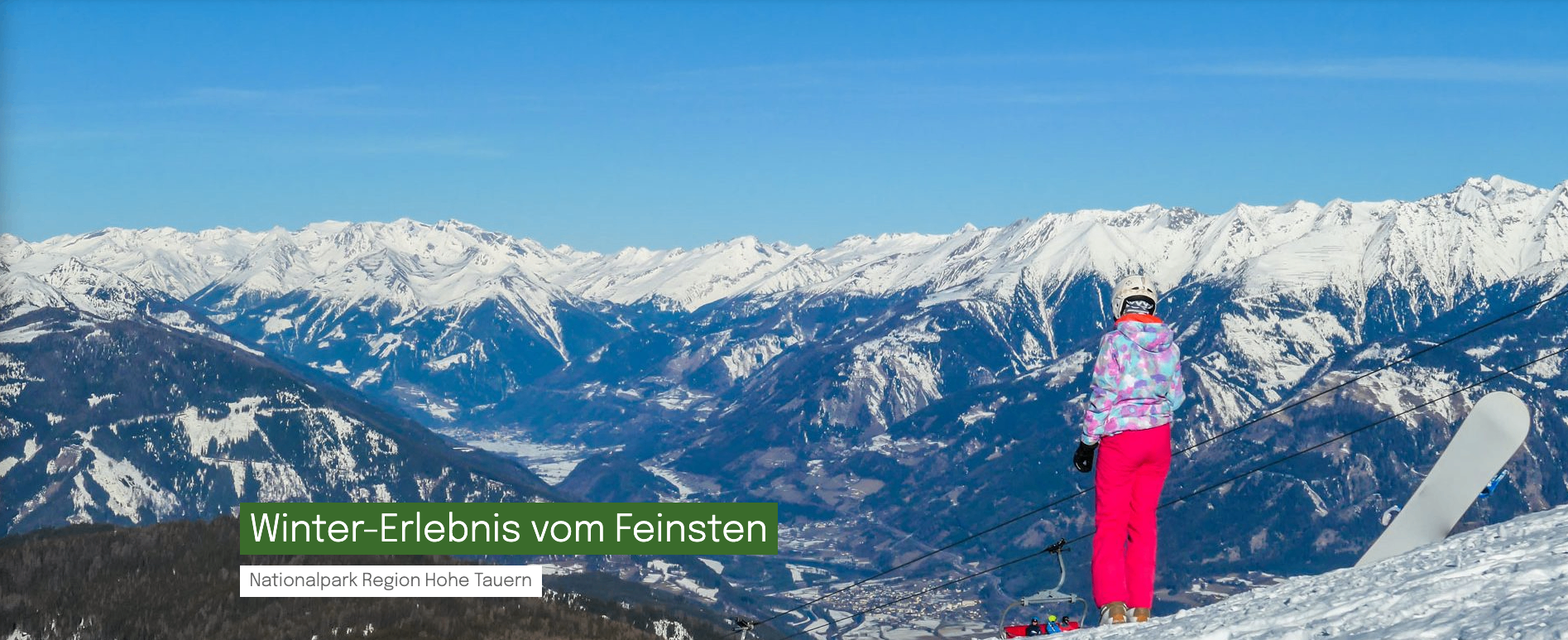 Ein Panoramablick auf schneebedeckte Berge mit einer Skilift im Vordergrund. Der Himmel ist klar und blau.