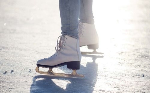 Eine Person in weißen Schlittschuhen fährt auf dem Eis, wobei blaue Jeans und hellblaue Hose sichtbar sind. Das Eis ist sauber und weiß, und die Schlittschuhe haben schwarze Klingen.