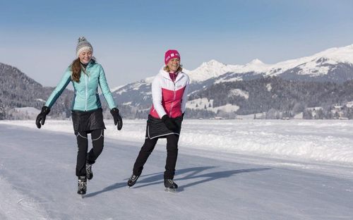 Zwei Frauen skaten auf einem gefrorenen See, umgeben von schneebedeckten Bergen unter einem klaren blauen Himmel.