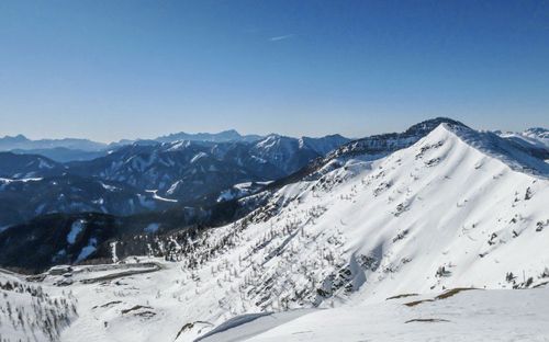 Ein Panoramablick auf eine verschneite Bergkette unter einem klaren blauen Himmel. Die Berghänge sind mit Schnee bedeckt und haben einige Bäume. In der Ferne gibt es eine kurvenreiche Straße.