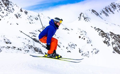 Ein Skifahrer in Blau und Orange ist in der Luft auf einem verschneiten Berg zu sehen, mit einem bewölkten Himmel im Hintergrund.