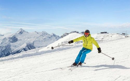 Ein Skifahrer in einer gelben Jacke fährt einen schneebedeckten Berg hinunter, mit Bergen im Hintergrund.