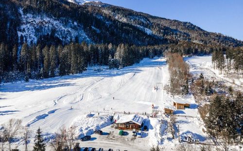 Ein Skigebiet mit verschneiten Hängen und Kiefern. Einige Menschen fahren die Piste hinunter. Die Skilifte befinden sich auf der rechten Seite. Gebäude und Autos sind rund um das Resort geparkt.