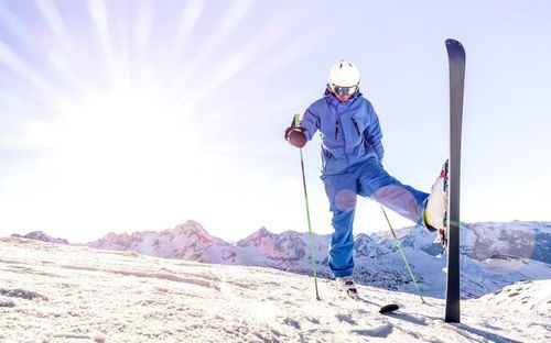 Ein Skifahrer in einer blauen Kleidung steht auf verschneiten Bergen, hält Skistöcke und einen Fuß in einem Skischuh.