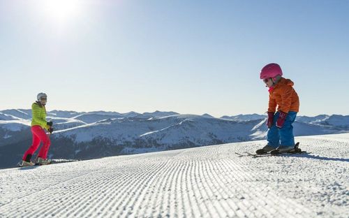 Zwei Skifahrer sind auf einem verschneiten Berg. Einer trägt einen rosa Helm und eine orange Jacke, der andere eine gelbe Jacke und rosa Hose. Sie stehen auf einem verschneiten Hang.