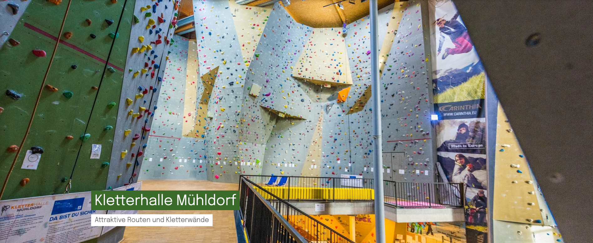 Die große Indoor-Kletterwand ist mit bunten Klettergriffen gefüllt. Unten befindet sich eine gelbe Plattform mit einem blauen Stuhl. Unten links ist der Name Muhldorf zu sehen.