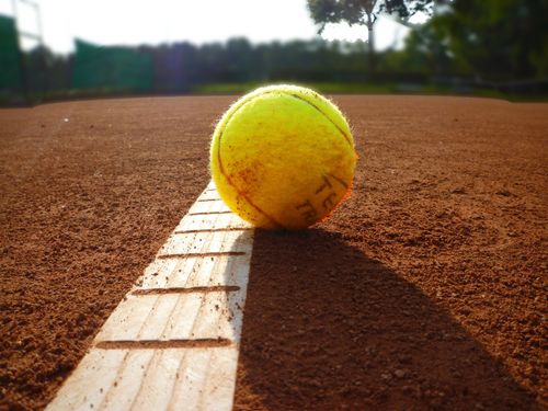Ein Tennisball liegt auf der Seitenlinie eines Tennisplatzes mit brauner Asche. Der Ball ist gelb mit schwarzen Schriftzeichen.