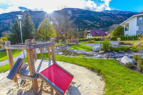 Ein Spielplatz mit Rutsche und Sandkasten. Im Hintergrund sind Gras, ein Haus und Berge zu sehen. Die Sonne scheint hell.