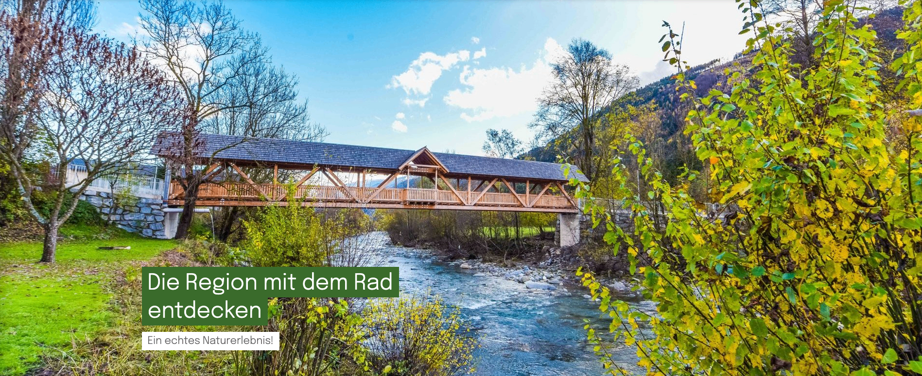 Eine Brücke überspannt einen Fluss, mit Felsen und Bäumen in der Nähe. Die Worte 'mit dem Rad' sind unten sichtbar.